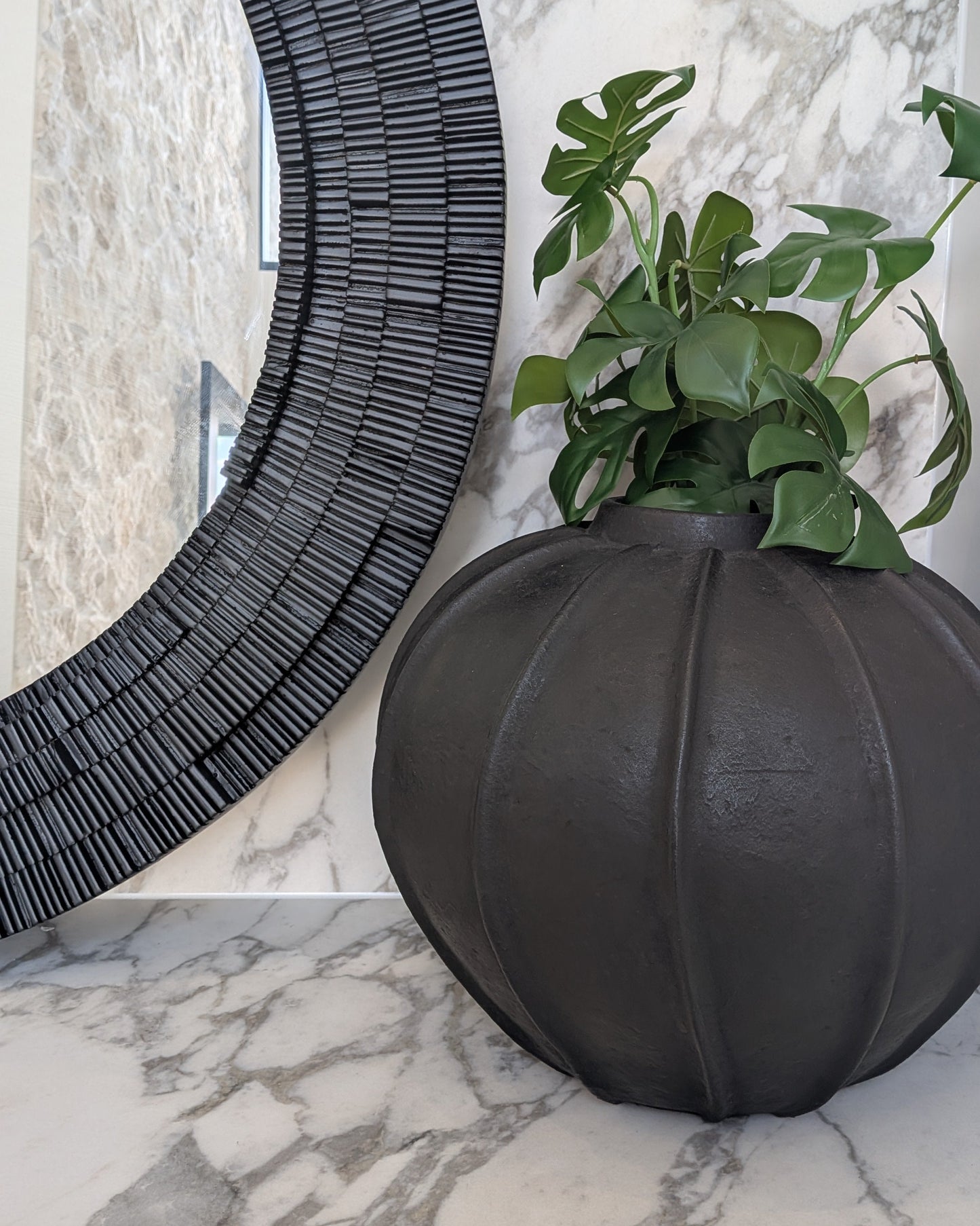 Decorative black vase with green plant on a marble surface next to a textured black round mirror.