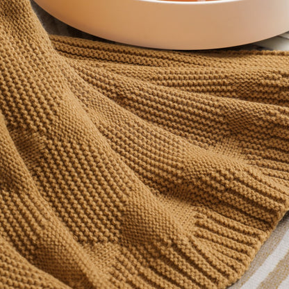 Brown knitted blanket draped over a surface with a blurred white object in the background