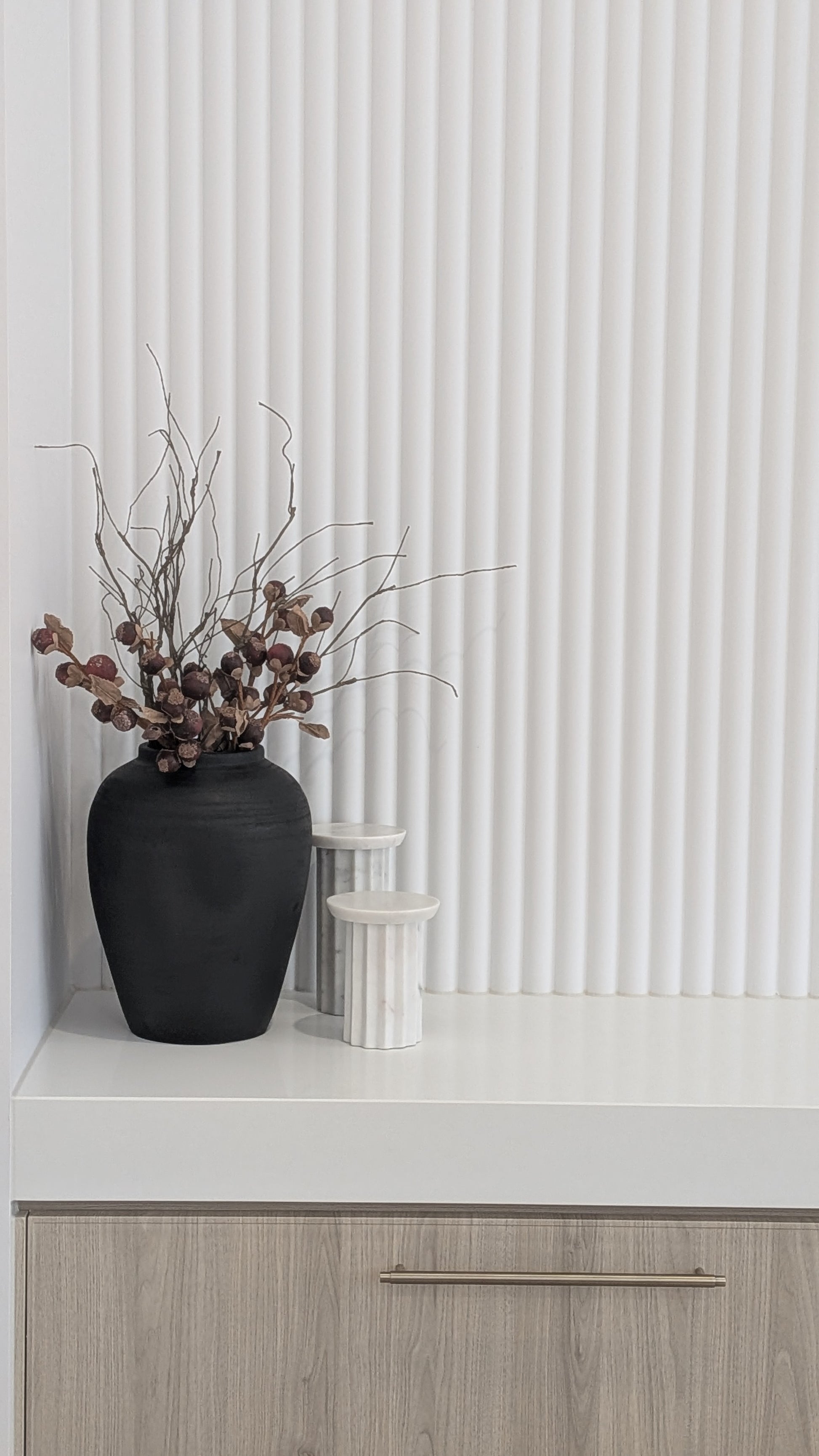 Black vase with dried branches on a white stone bench against a fluted white wall.