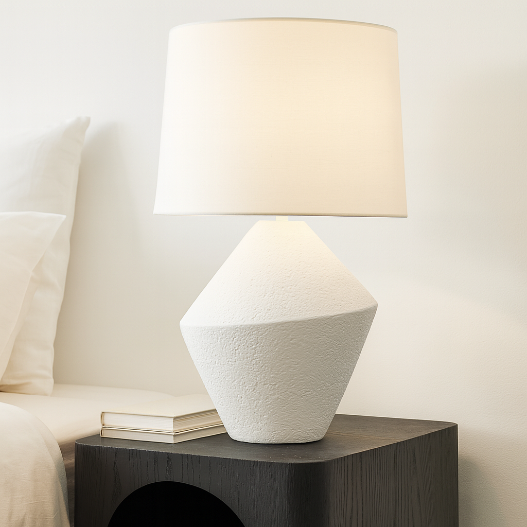 Table lamp with textured base and white shade on a nightstand next to a bed.
