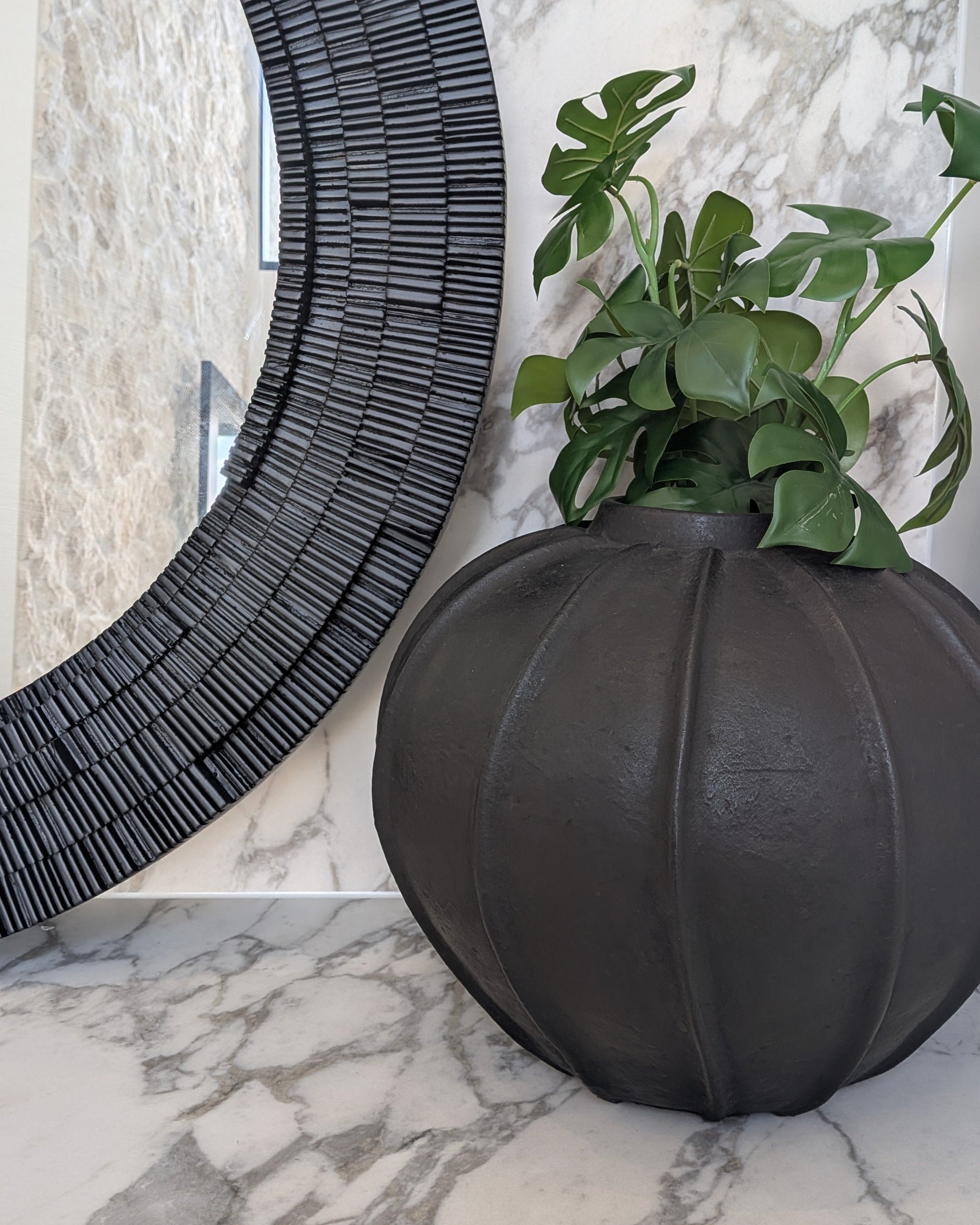 Decorative black vase with green plant on a marble surface next to a textured black round mirror.