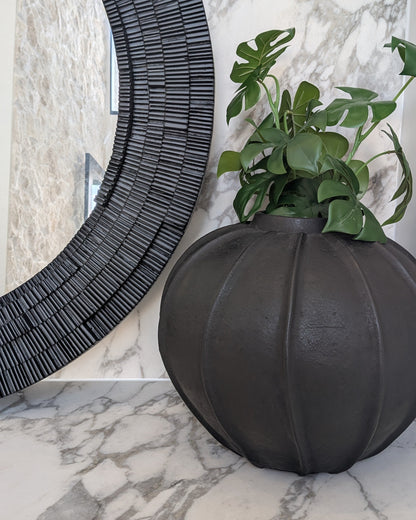 Decorative black vase with green plant on a marble surface next to a textured black round mirror.