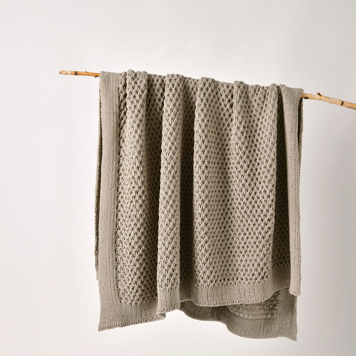 Knitted blanket with a textured pattern draped over a wooden stick on a light background